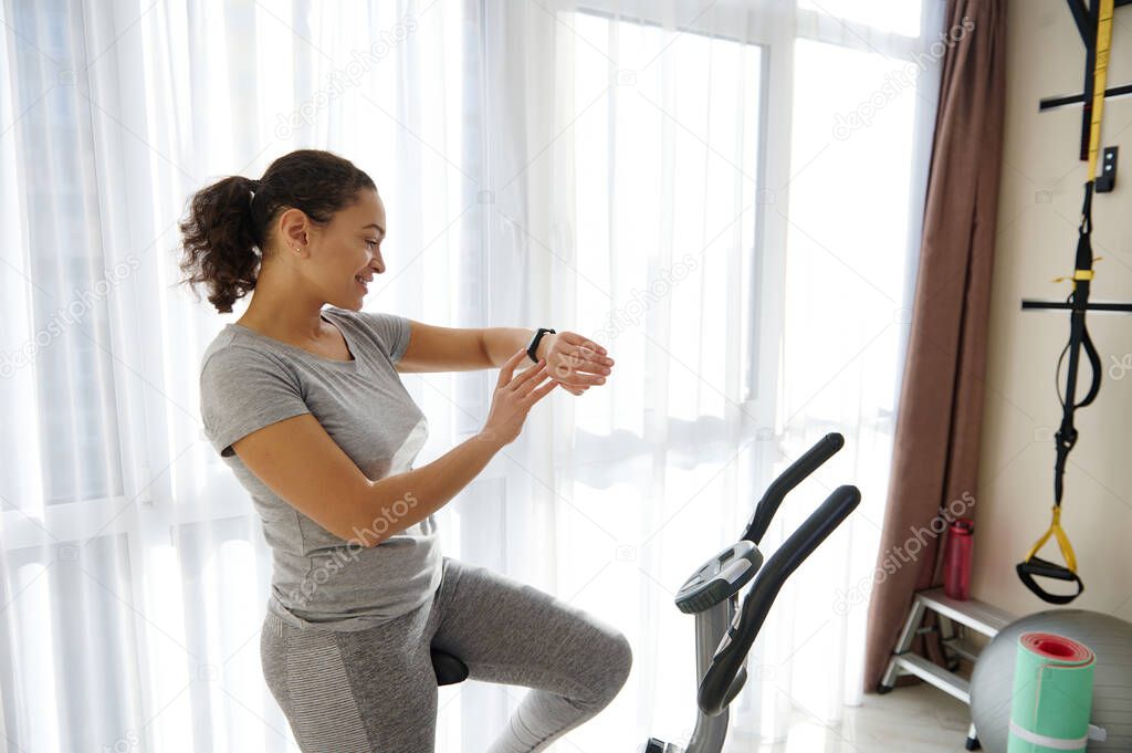 Happy woman checking her fitness tracker and heart rate while standing on a spin bike after cardio workout. Fit woman enjoying workout at home
