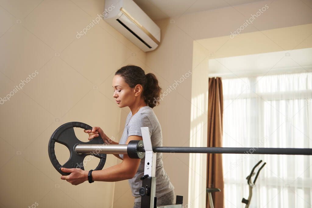 A young fit woman puts a metal disc on an Olympic barbell during bodybuilding workout. Exercising at home.