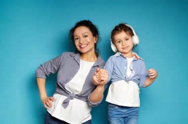 Çekici bir kadın kameranın önünde mavi bir arka planda dişlek bir gülümsemeyle poz veriyor ve küçük kızı kulaklık takıyor.