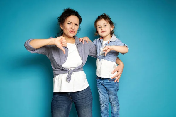 Beautiful mom and daughter equally dressed showing thumbs down and expressing dissatisfaction and displeasure while posing to camera on blue background with copy space