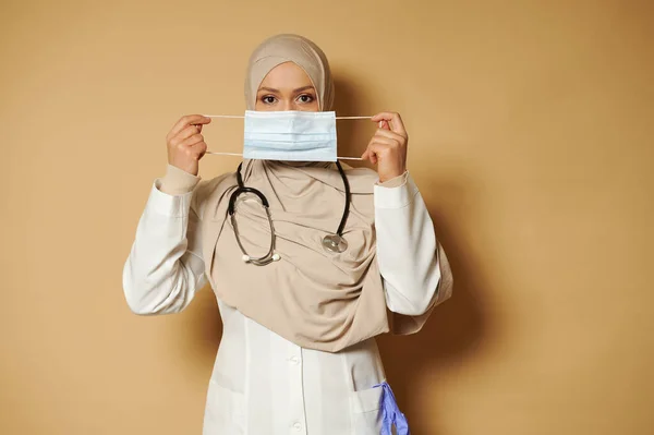 Muslim doctor with in hijab holding and wearing medical mask standing on beige background eith copy space