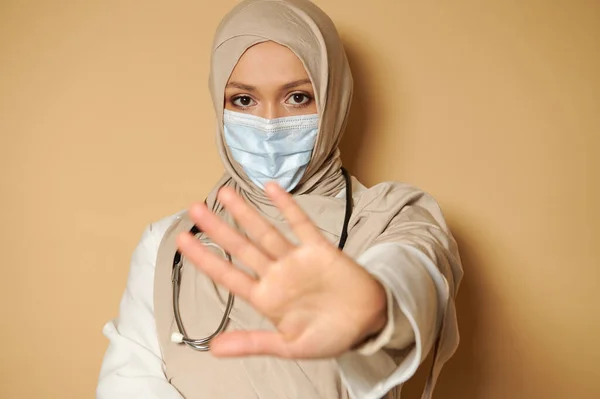 Portrait of muslim doctor woman in hijab and medical mask gesturing STOP with her hand. Beige background, copy space.