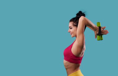 Güzel ve narin atletik kadın, mavi bir arka planda yan yana dururken kol ve kaslarını halterlerle şişiriyor.