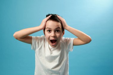 Shocked boy screams and laughs with his hands on his temples, expressing shock and surprise. Blue background, copy space clipart