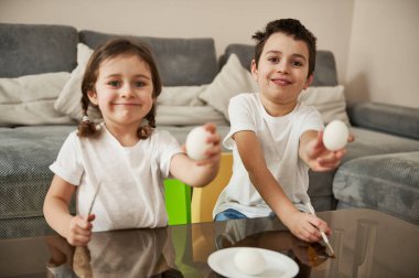 Paskalya yumurtalarını süslemek için ellerinde boya fırçası olan beyaz haşlanmış yumurtaları gösteren sevimli çocuklar. Paskalya konsepti