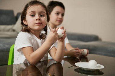 Güzel kız bir okul çocuğunun yanında oturuyor, elinde fırça tutuyor ve Paskalya yumurtalarını boyamadan önce kameraya gülümsüyor..