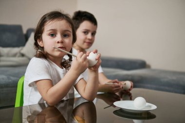 Güzel bir kız, elinde boya fırçası olan bir çocuğun yanında oturuyor ve Paskalya yumurtalarını boyamadan önce kameraya bakıyor..