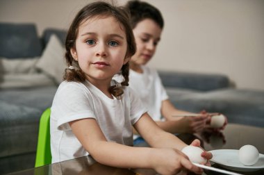 Güzel kız bir okul çocuğunun yanında oturuyor, elinde fırça tutuyor ve Paskalya yumurtalarını boyamadan önce kameraya gülümsüyor..