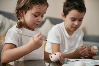 Beyaz tişörtlü, kardeşinin yanında oturan sevimli küçük kız Paskalya yumurtalarını süslemek için boya fırçası kullanıyor. Geleneksel dini olayı kutlamaya hazır mısın?.