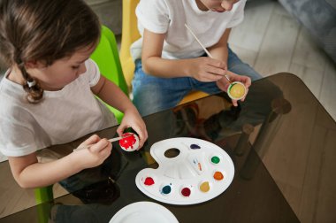 Paskalya yumurtalarını evde süsleyen iki sevimli çocuğun yüksek açılı görüntüsü. Sanatsal etkinlik, dekorasyon, Paskalya kutlaması konsepti