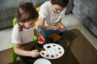 Paskalya yumurtalarını evde süsleyen iki sevimli çocuğun yüksek açılı görüntüsü. Sanatsal etkinlik, dekorasyon, Paskalya kutlaması konsepti