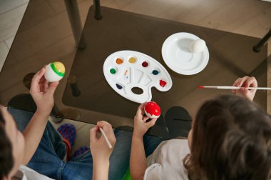 Paskalya yumurtalarını süsleyen iki çocuk. Tabağında boya ve cam bir masanın üstünde yumurta olan bir palet. Yüksek açı görünümü.