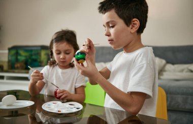 Erkek ve kız aynı giyinip Paskalya yumurtalarını boyayla süslüyor oturma odasındaki masada oturuyor.