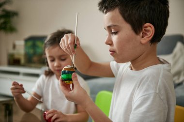 Okul çocuğu evde küçük kız kardeşinin yanında otururken bir fırça ve renkli boyalarla Paskalya yumurtasını süslemeye odaklandı..