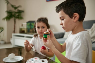 Okul çocuğu evde küçük kız kardeşinin yanında otururken bir fırça ve renkli boyalarla Paskalya yumurtasını süslemeye odaklandı..