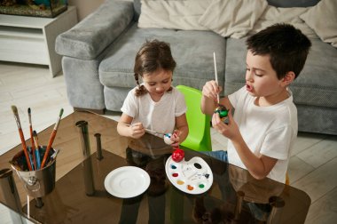 Mutlu çocuklar bir masada oturuyor, Paskalya için yumurtaları renkli boyalarla süslüyorlar.