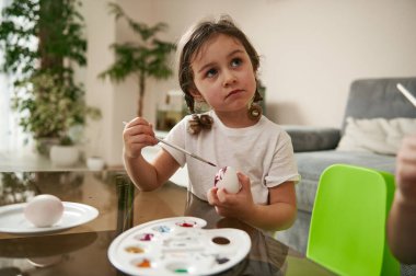 Saçları örgülü sevimli bir anaokulu öğrencisi Paskalya yumurtasını süslüyor, masada oturuyor ve başka tarafa bakıyor. Paskalya.