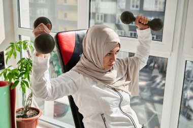 Türbanlı güzel bir Arap kadının portresi basın bankında oturuyor ve evde demir halterlerle egzersiz yapıyor.