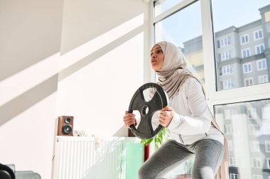 Başlıklı, tesettürlü, elinde metal bir diskle ağırlıklı çömelme hareketleri yapan Arap Müslüman kadın. Ev ödevi kavramı..