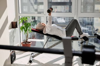 Kafası tesettüre bulanmış, spor salonunda halterli göğüs presi yapan, arka planda geniş pencereler olan kararlı Arap Müslüman kadının yan görüntüsü