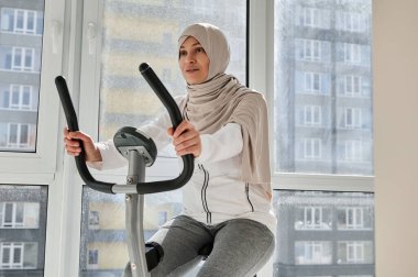 Güzel, güneşli bir günde evde bisikletle egzersiz yapan çekici Müslüman kadın. Kardiyo çalışması. Evde fitness