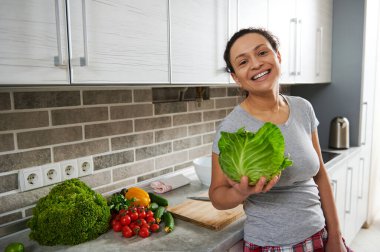 Elinde lahana tutan ve dişlek bir gülümsemeyle gülümseyen, kameraya bakan, evdeki mutfağın arka planında duran genç bir kadın.