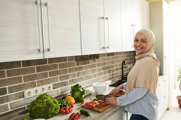 Kafası tesettüre bulanmış çekici Arap kadın kameraya bakarken malzemeleri kesiyor ve mutfakta sebze salatası hazırlıyor. Vegan gıda konsepti. Sağlıklı beslenme. Evde yemek pişirmek..