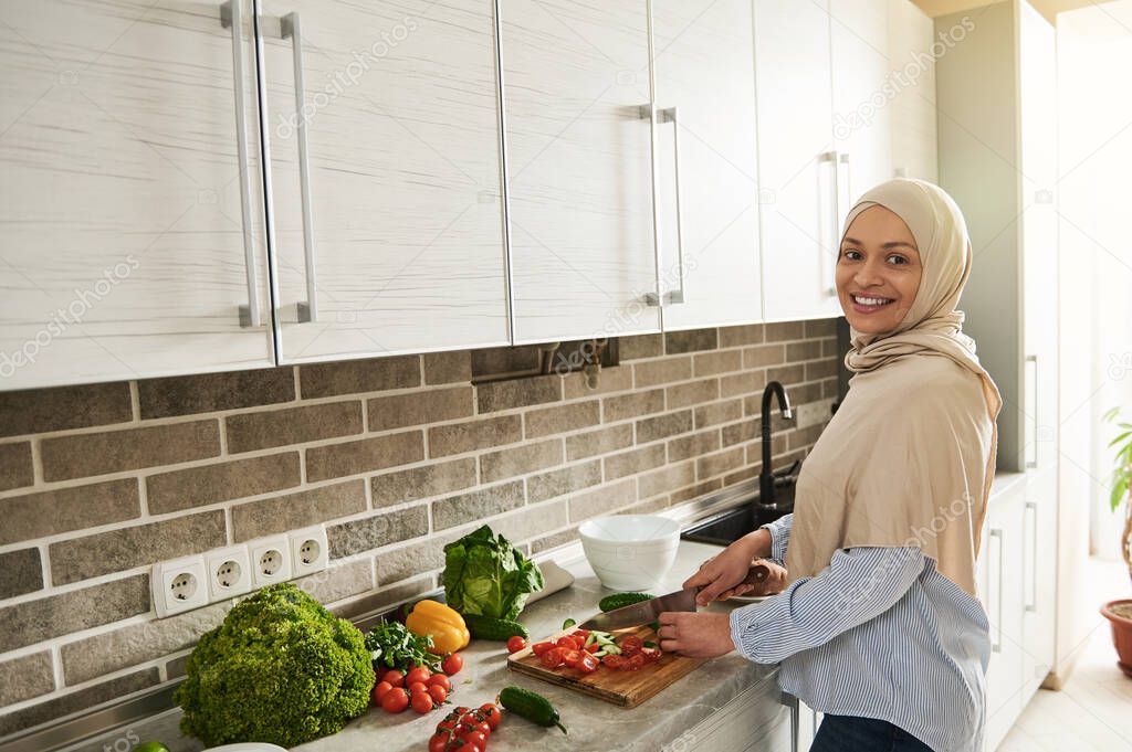 Attractive Arab Muslim woman with covered head in hijab looks at camera while cutting ingredients and preparing vegetable salad in the kitchen. Vegan food concept. Healthy eating. Cooking at home.