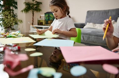 Kardeşinin yanında oturan ve renkli bir kağıda resim çizen küçük bir anaokulu kızına yan bakış.