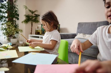 Kardeşinin yanında oturan ve renkli bir kağıda resim çizen küçük bir anaokulu kızına yan bakış. Evde yaratıcılık dersi