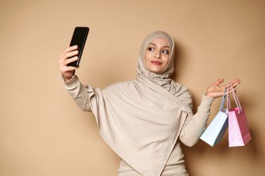 Bir elinde mavi ve pembe çantalar, diğer elinde akıllı telefon tutan çekici Müslüman kadın selfie çekiyor. Alışveriş konsepti, etkinlikler için hediye.