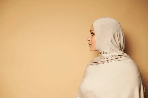 Side portrait of young Arab Muslim woman in strict religious clothes and head covered in hijab looking to the side against beige background with copy space