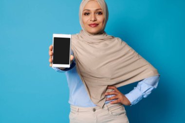 Arap Müslüman kadının elindeki cep telefonuna odaklanın. Başörtülü genç bir kadın elinde akıllı bir telefon ve mavi arkaplan üzerinde poz metin için boşluk ile