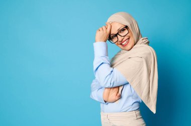 Bej tesettürlü çekici Müslüman kadın tesettürlü tesettürlü tesettürlü tesettürlü ve mavi arka planda fotokopi aletiyle kameranın yanında duruyor.
