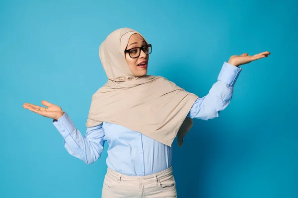 ヒジャーブのアラブ人女性は腕と手のひらを伸ばしてポーズをとり テキストのためのスペースを持つ青の背景に提示 — ストック写真