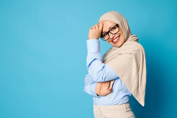 魅力的なイスラム教徒の女性とともにカバー頭でベージュヒジャーブ笑顔とおもちゃの笑顔とスタンド側にカメラの上に青の背景とともにコピースペース — ストック写真