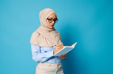 Müslüman bir kadın gözlük takıyor ve kafasında beyaz bir kitap yazılı sıkı bir kıyafet giyiyor. Mavi arka planda, kopya alanı ile izole edilmiş.