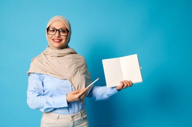 Başörtüsü içinde gülümseyen Müslüman kadın boş bir kitap tutuyor ve onu mavi arka planda izole edilmiş kalemle işaret ediyor.
