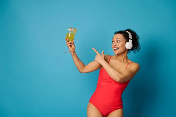 Charming woman with healthy and aesthetic body wearing red swimsuit and headphones looking and pointing at an exotic cocktail in her hand, isolated over blue background with copy space