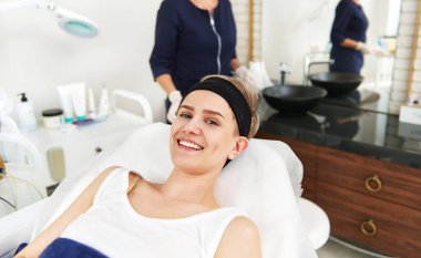 Güzel bir gülümsemesi olan güzel bir kadın. Masaj masasında dinleniyor. Arka planda tanınmayan bir doktor kozmetik uzmanı.