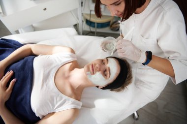 Güzellik uzmanının kaplıca kliniğindeki güzel bir kadının yüzüne aljinik maske taktığı yüksek açılı bir manzara. Profesyonel cilt bakımı kavramı