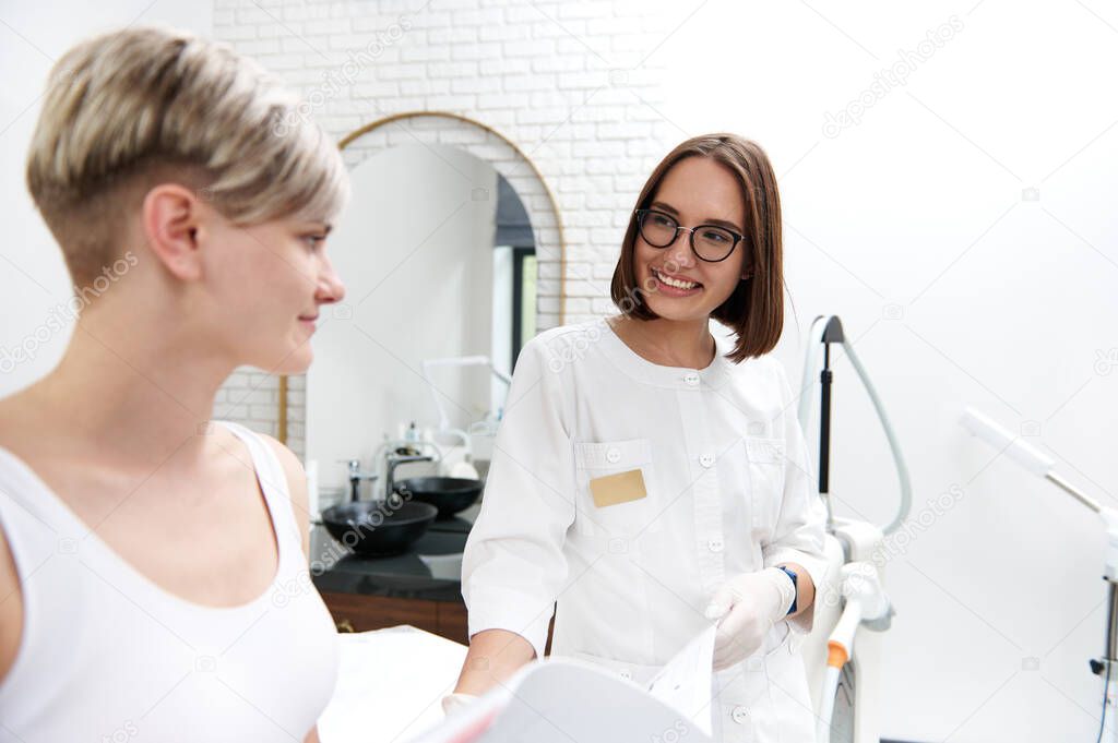 Smiling cosmetologist showing a catalog of prices for cosmetic procedures to a young client g during a consultation at a beauty spa clinic