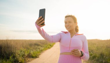 Güzel bir sporcu kadın elinde akıllı bir telefonla ıssız bir bozkırda duruyor ve güzel gülücükler açık havada kardiyo çalışmasının keyfini çıkarıyor. Spor, spor ve eğitim kavramları.