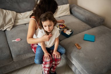 Yüksek görüş açısı. Annem kızının saçını tarıyor ve güzel bir saç stili için saç tokası seçiyor. Mutlu çocukluk ve annelik kavramı. Aşk, ilgi, şefkat.