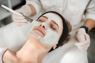 Bir güzellik uzmanının elinin yakın plan portresi fırçalı, kusursuz bir cilde sahip genç bir kadının yüzüne güzellik maskesi takıyor. Modern güzellik salonlarında profesyonel cilt bakımı konsepti