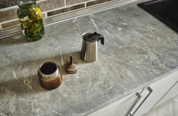 High angle view f a disassembled steel geyser coffee maker next to a glass bottle of lemon-mint water on the kitchen countertop.