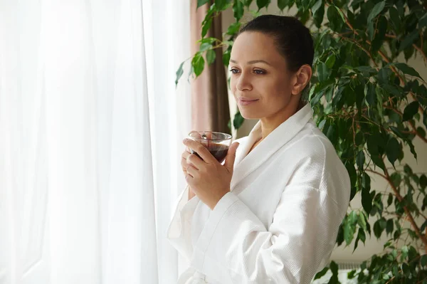 A serene mature woman in a white robe stands with a cup of coffee in her hands against the background of a houseplant and enjoys the view outside the window through transparent white curtains. Morning routine concepts