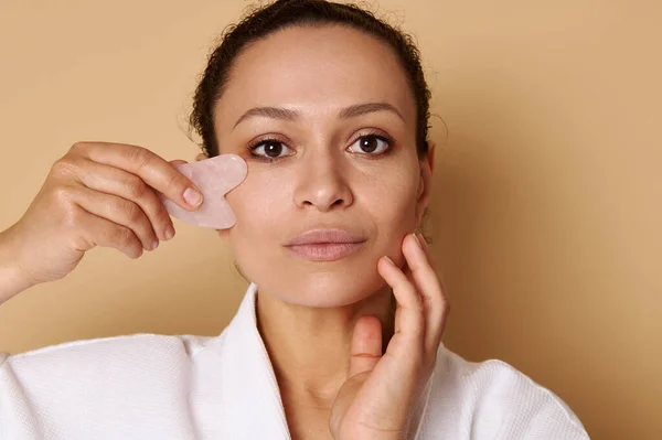 Lenfatik drenaj yüz masajı için gua-sha kullanan melez kadının güzel portresi. Spa ve cilt bakımı kavramları.