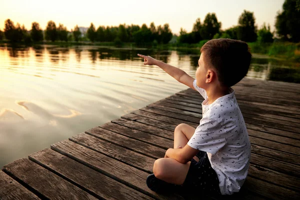 stock image Silhouette of a serene child sitting on the wooden pier and enjoying heat summer evening at the lake at sunset. Peace and tranquility on nature background