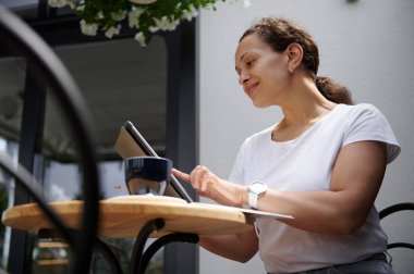 Gündelik giysiler içindeki çekici olgun kadın yaz terasında bir masada oturur ve dijital tablet üzerinde çalışır. Serbest meslek, iş ve açık hava iş kavramları